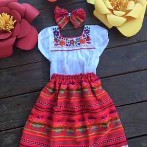 May include: A white embroidered blouse with a red and white skirt. The skirt has a colorful geometric pattern and a white lace trim. The outfit is decorated with paper flowers in red, orange, and yellow.