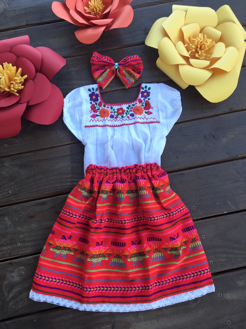 May include: A white embroidered blouse with a red and white skirt. The skirt has a colorful geometric pattern and a white lace trim. The outfit is decorated with paper flowers in red, orange, and yellow.