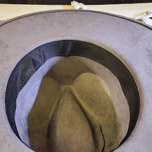 May include: A light gray cowboy hat with a dark gray band and a black interior. The hat is made of a soft, suede-like material. The brim curves upwards, and the crown has a classic teardrop shape.