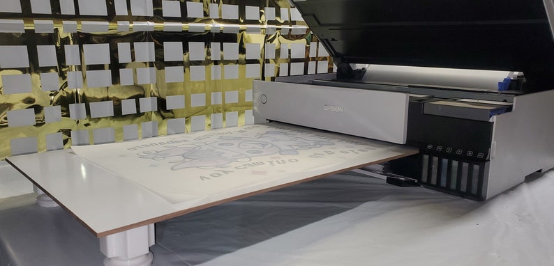 May include: A white printer with an open lid, printing on a white sheet of paper with a black and white design. The printer has a row of colored ink cartridges on the right side.