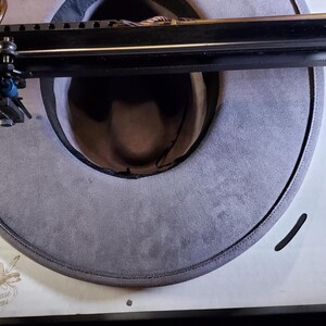 May include: A gray cowboy hat is positioned on a white surface, with a laser cutting machine above it. The hat's brim is visible, and the text "Showcase Brims" is printed on the surface.