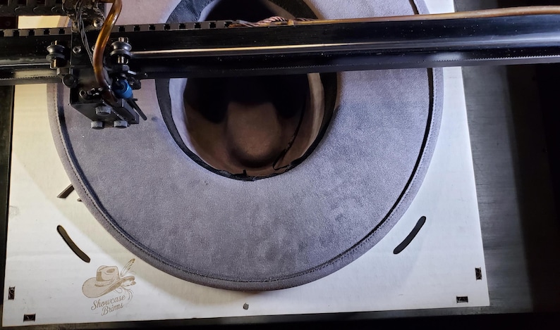 May include: A gray cowboy hat is positioned on a white surface, with a laser cutting machine above it. The hat's brim is visible, and the text "Showcase Brims" is printed on the surface.