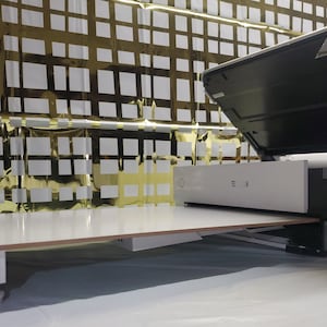May include: A white flatbed scanner with a black lid open. The scanner is on a white surface with a gold and white patterned background.