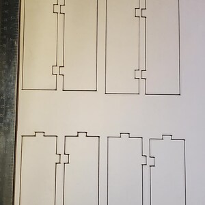 May include: A white sheet of paper with eight rectangular shapes drawn on it. Each rectangle has a series of notches and tabs, likely for interlocking or assembly. A metal ruler is visible on the left side.