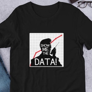 Pode incluir: Camiseta preta com um gráfico branco de uma pessoa segurando um microfone e um gráfico de linha vermelha. O texto no gráfico diz "Show Me The Data!"