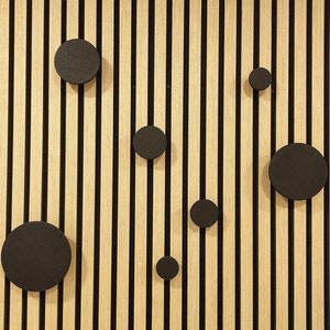 Op de afbeelding: Een muur met verticale houten latten die in een lichtbruine kleur zijn geverfd. De latten worden gescheiden door zwarte lijnen. Er zijn zes zwarte cirkels van verschillende grootte aan de muur bevestigd.