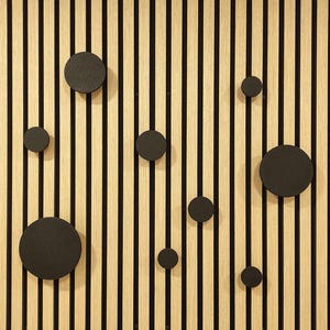 Puede incluir: Un panel de pared de madera con rayas negras verticales y vetas de madera de color marr&oacute;n claro. Varios ganchos de pared negros y circulares de diferentes tama&ntilde;os est&aacute;n adheridos al panel, creando un dise&ntilde;o decorativo y funcional.