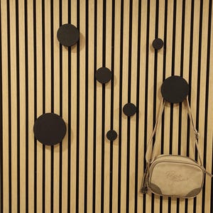 Puede incluir: Un panel de pared de madera con rayas negras verticales y listones de color madera natural. Varios ganchos circulares negros est&aacute;n montados en el panel. Una bandolera beige con el texto "Pepe Jeans" cuelga de uno de los ganchos.