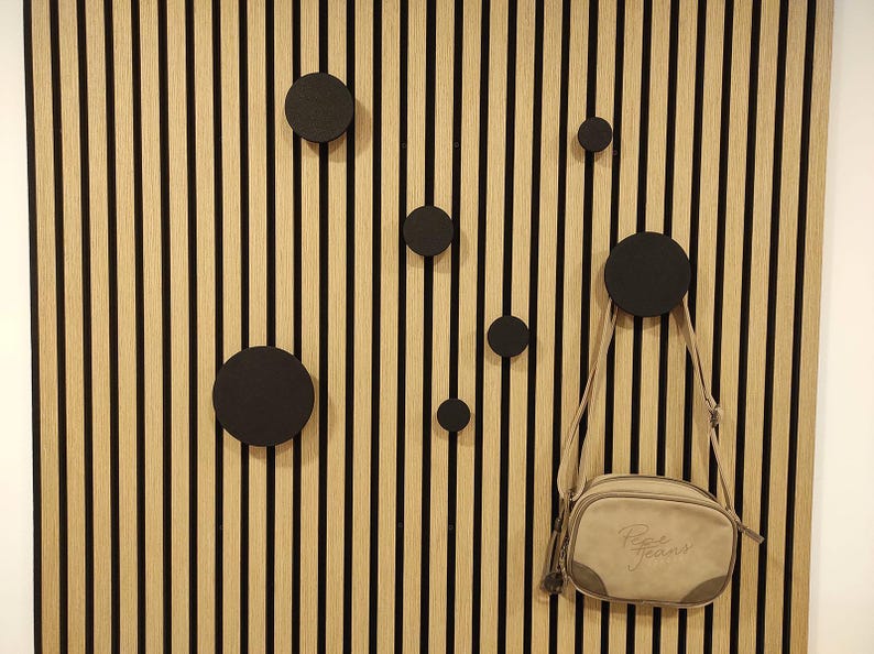 Puede incluir: Un panel de pared de madera con rayas negras verticales y listones de color madera natural. Varios ganchos circulares negros est&aacute;n montados en el panel. Una bandolera beige con el texto "Pepe Jeans" cuelga de uno de los ganchos.