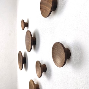 Könnte beinhalten: Eine Ansammlung von Wandhaken aus Holz in verschiedenen Größen und Formen, an einer weißen Wand befestigt. Die Haken sind aus warm getöntem Holz gefertigt, das die natürliche Maserung zeigt. Das Design ist minimalistisch und modern und für verschiedene Einrichtungsstile geeignet.