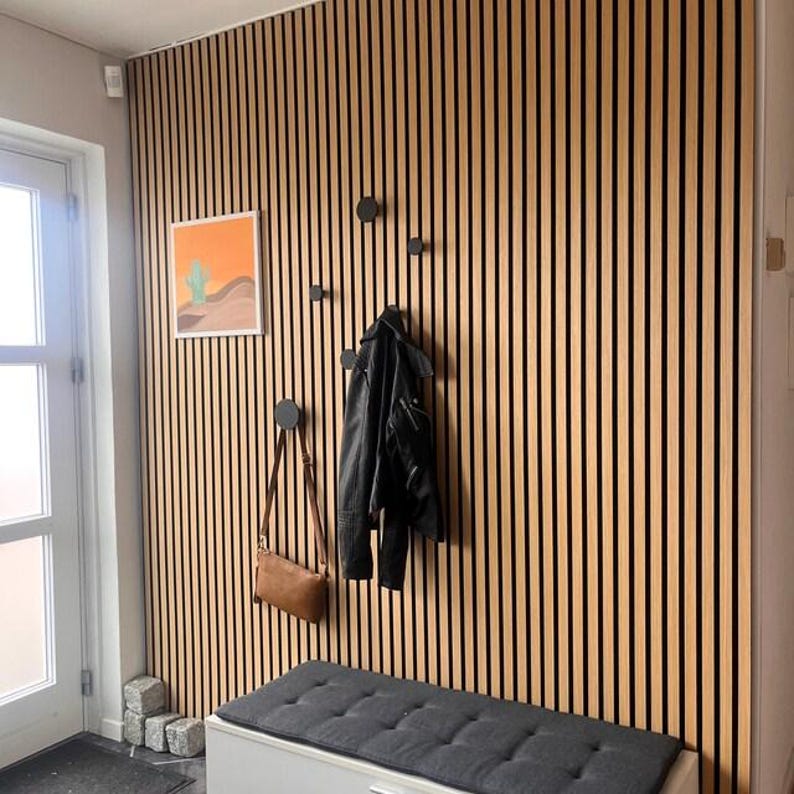 Puede incluir: Entrada moderna con una pared de listones de madera, con una obra de arte enmarcada de un paisaje des&eacute;rtico, percheros negros, una chaqueta negra y un bolso marr&oacute;n. Debajo hay un banco gris con un coj&iacute;n acolchado.