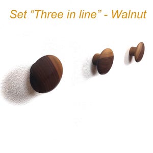 May include: Three walnut wood knobs mounted on a white wall. The knobs are oval-shaped and vary in size, arranged in a line. The text "Set 'Three in line' - Walnut" is in the upper left corner.