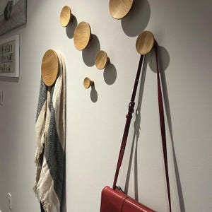 Könnte beinhalten: An einer weißen Wand sind Holz-Wandhaken in verschiedenen Größen angebracht. Eine rote Handtasche hängt an einem der Haken, und ein Schal ist über einen anderen gehängt. Die Haken haben eine natürliche Holzmaserung.
