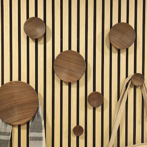 May include: A wall with wooden slats in a light brown color with black lines running vertically. There are six round wooden hooks attached to the wall. One hook has a gray and white scarf hanging from it. A brown leather bag is hanging from another hook.