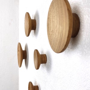 Könnte beinhalten: Eine Ansammlung von Wandhaken aus Holz in verschiedenen Größen, an einer weißen Wand befestigt. Die Haken sind aus hellem Holz gefertigt und haben ein schlichtes, rundes Design. Der größte Haken hat einen Durchmesser von etwa 10 cm.