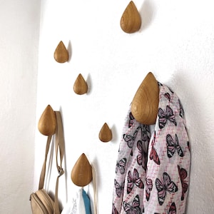 Peut inclure: Des crochets muraux en bois en forme de goutte d'eau de différentes tailles sont montés sur un mur blanc. Un sac à main beige, un sac blanc et bleu et une écharpe à motif de papillons roses sont suspendus aux crochets. Les crochets sont en bois clair.