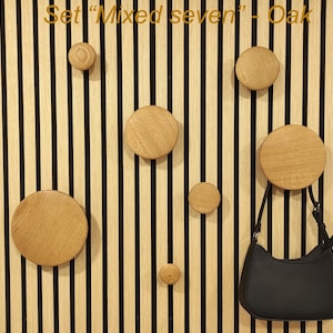 May include: A set of seven wooden wall hooks in varying sizes, mounted on a vertical striped wooden panel. A black handbag hangs from one of the hooks. The text "Set Mixed seven - Oak" is at the top.