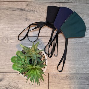 May include: Three face masks with black elastic straps. The masks are green, dark blue, and black. The masks are lying on a wood surface with a potted succulent plant.