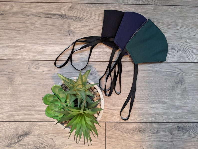 May include: Three face masks with black elastic straps. The masks are green, dark blue, and black. The masks are lying on a wood surface with a potted succulent plant.