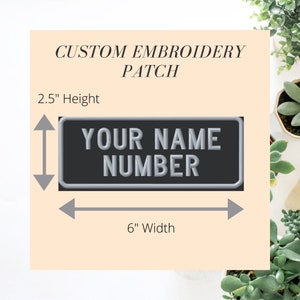 May include: Custom embroidery patch, 15.2 cm wide and 6.3 cm high, with the text "YOUR NAME NUMBER" in a 3D style. The patch has a black background and a silver border. The words "CUSTOM EMBROIDERY PATCH" are above the main text.