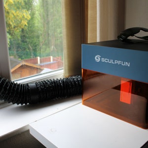 May include: A blue and orange 3D printer with the logo "Sculpfun" on the side. A black flexible hose is connected to the printer. The printer is on a white desk next to a window.