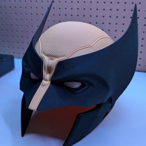 May include: A black and tan Wolverine mask, with the character's distinctive pointed ears and eye openings. The mask has a textured surface, possibly plastic. The design is a replica of the comic book character, a popular superhero.