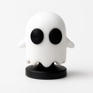 May include: A white ghost-shaped figurine with two large, black, textured eyes. The ghost has small, rounded arms and a scalloped bottom edge. It is mounted on a black, circular base, creating a whimsical, decorative object.