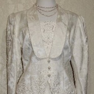 May include: A white, long-sleeved,  brocade jacket and skirt set with a lace trim. The jacket has a high collar and a fitted waist. The skirt is a-line and falls to the knee.