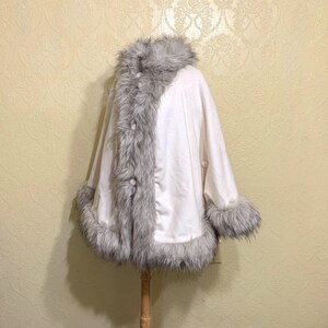 May include: A white faux fur cape with a gray faux fur trim around the collar, cuffs, and down the front. The cape has a button closure.