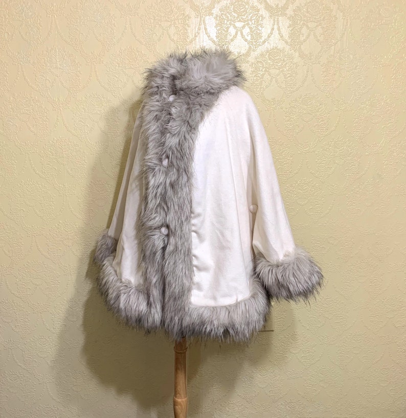 May include: A white faux fur cape with a gray faux fur trim around the collar, cuffs, and down the front. The cape has a button closure.