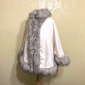 May include: A white faux fur poncho with a gray faux fur trim around the collar, sleeves, and hem. The poncho has a button closure.