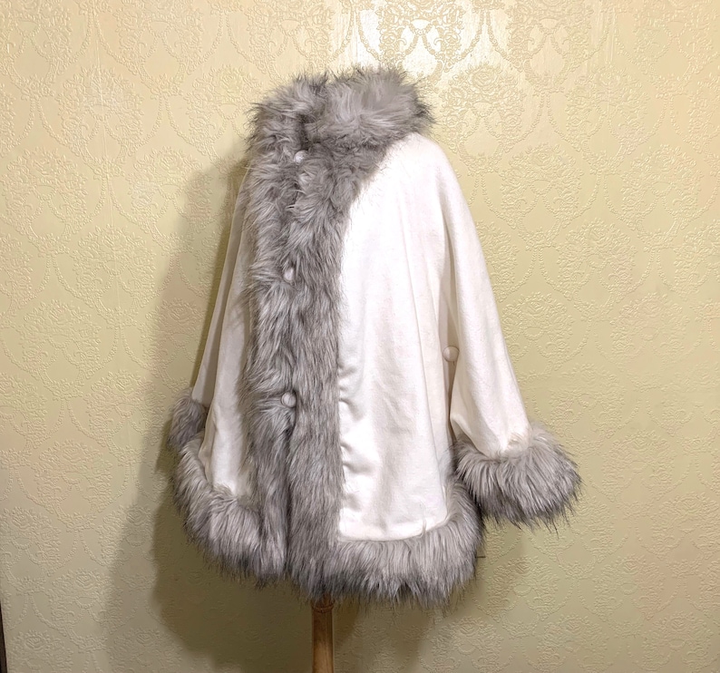 May include: A white faux fur poncho with a gray faux fur trim around the collar, sleeves, and hem. The poncho has a button closure.