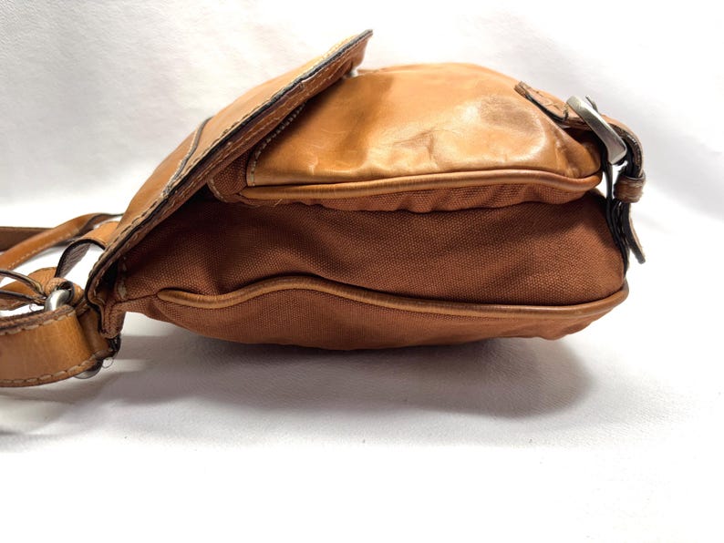 Pu&ograve; includere: Una borsa a tracolla marrone con chiusura a patta. La borsa presenta una combinazione di pelle e tela. La pelle &egrave; di colore marrone chiaro e la tela &egrave; di un marrone pi&ugrave; scuro. La borsa ha una fibbia argentata e una lunga tracolla.