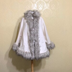 May include: A white faux fur poncho with a gray faux fur trim around the collar, hem, and sleeves. The poncho has three white buttons down the front.