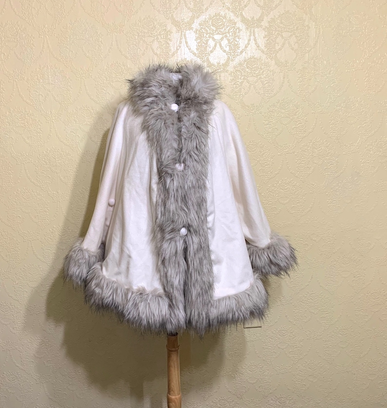 May include: A white faux fur poncho with a gray faux fur trim around the collar, hem, and sleeves. The poncho has three white buttons down the front.