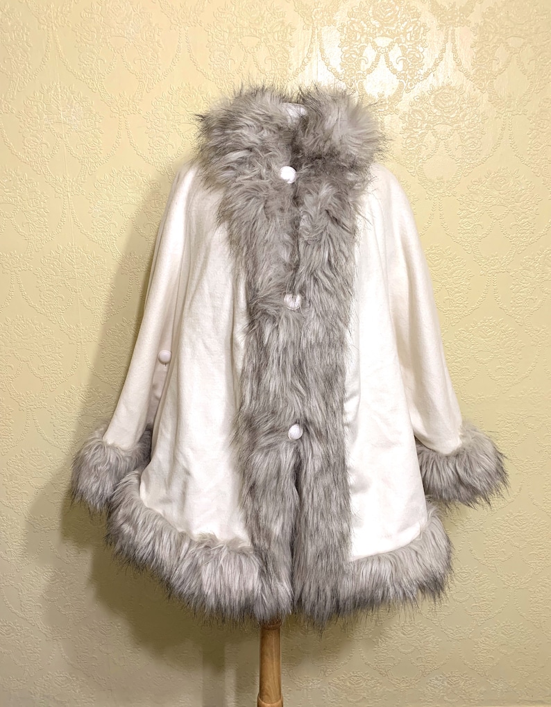 May include: A white faux fur cape with a gray fur trim around the collar, hem, and down the front. The cape has white buttons down the front.