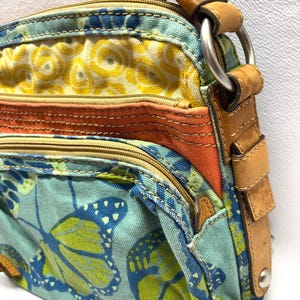 Pu&ograve; includere: Borsa a tracolla colorata con motivo floreale e a farfalle. La borsa presenta un mix di tessuti gialli, arancioni e verde acqua, con dettagli in pelle e un anello di metallo per la tracolla.