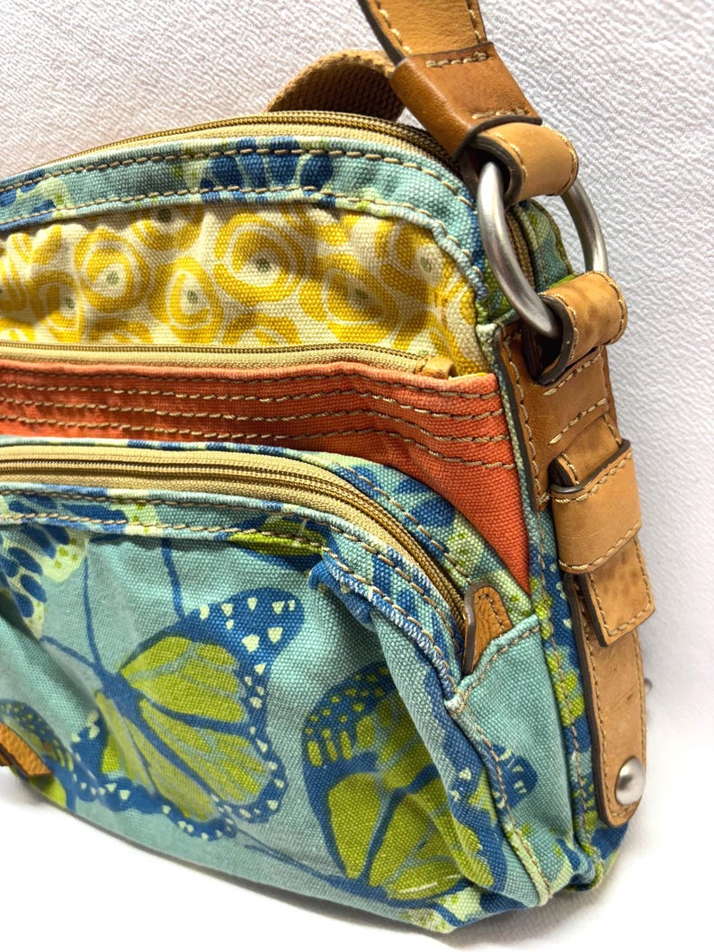 Pu&ograve; includere: Borsa a tracolla colorata con motivo floreale e a farfalle. La borsa presenta un mix di tessuti gialli, arancioni e verde acqua, con dettagli in pelle e un anello di metallo per la tracolla.