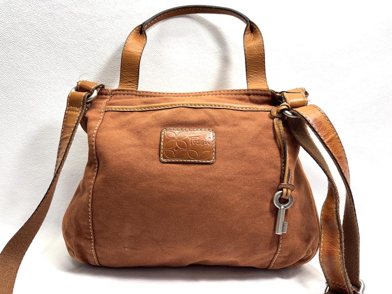 Pu&ograve; includere: Borsa a tracolla Fossil marrone con manico superiore e tracolla regolabile. La borsa presenta un ciondolo a forma di chiave e una toppa in pelle con il logo Fossil. La borsa &egrave; realizzata in materiale simile alla tela.