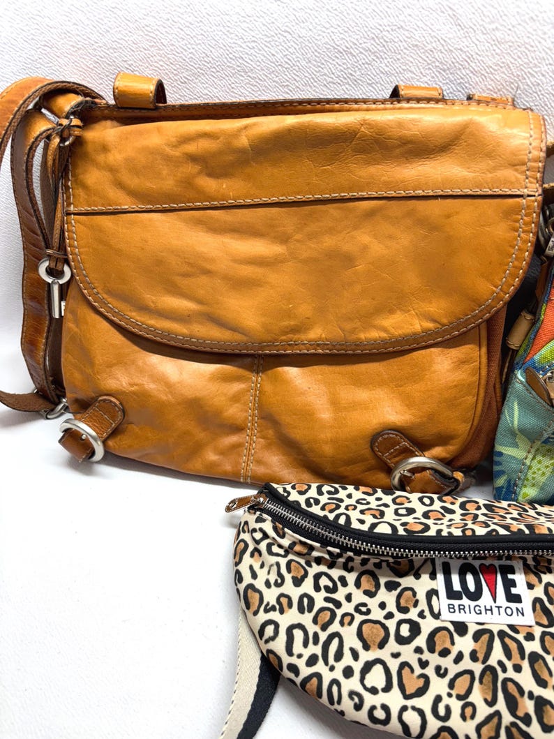 Pu&ograve; includere: Una borsa a tracolla in pelle marrone con patta curva e tracolla regolabile. La borsa ha finiture in metallo e un ciondolo a forma di chiave. &Egrave; visibile anche un marsupio con stampa leopardata con la scritta "LOVE BRIGHTON".