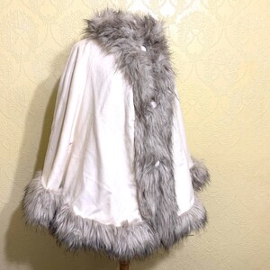 May include: A white faux fur cape with a gray faux fur trim around the collar, hem, and cuffs. The cape has a button closure at the front.