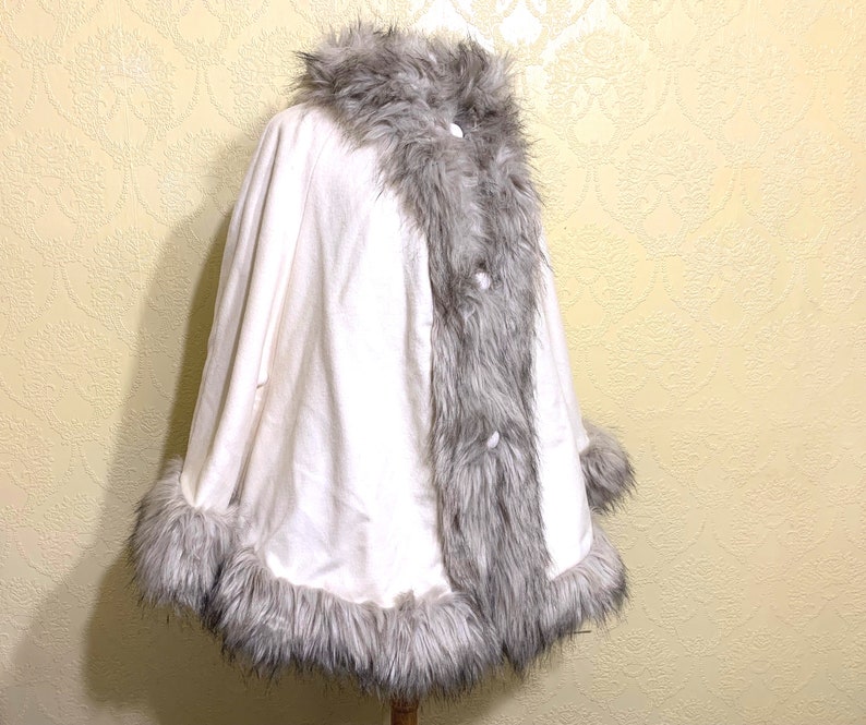 May include: A white faux fur cape with a gray faux fur trim around the collar, hem, and cuffs. The cape has a button closure at the front.