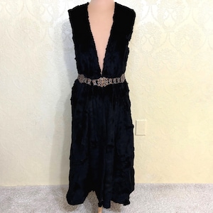May include: A long black faux fur vest with a deep V-neckline and a decorative belt. The vest has a flowing, layered design and is displayed on a mannequin.
