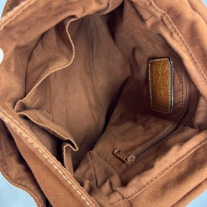 Pu&ograve; includere: Vista interna di una borsa tote in tela marrone con pi&ugrave; tasche interne e uno scomparto con cerniera. La borsa presenta cuciture bianche e un'etichetta in pelle con il nome del marchio. La fodera interna &egrave; di colore marrone abbinato.