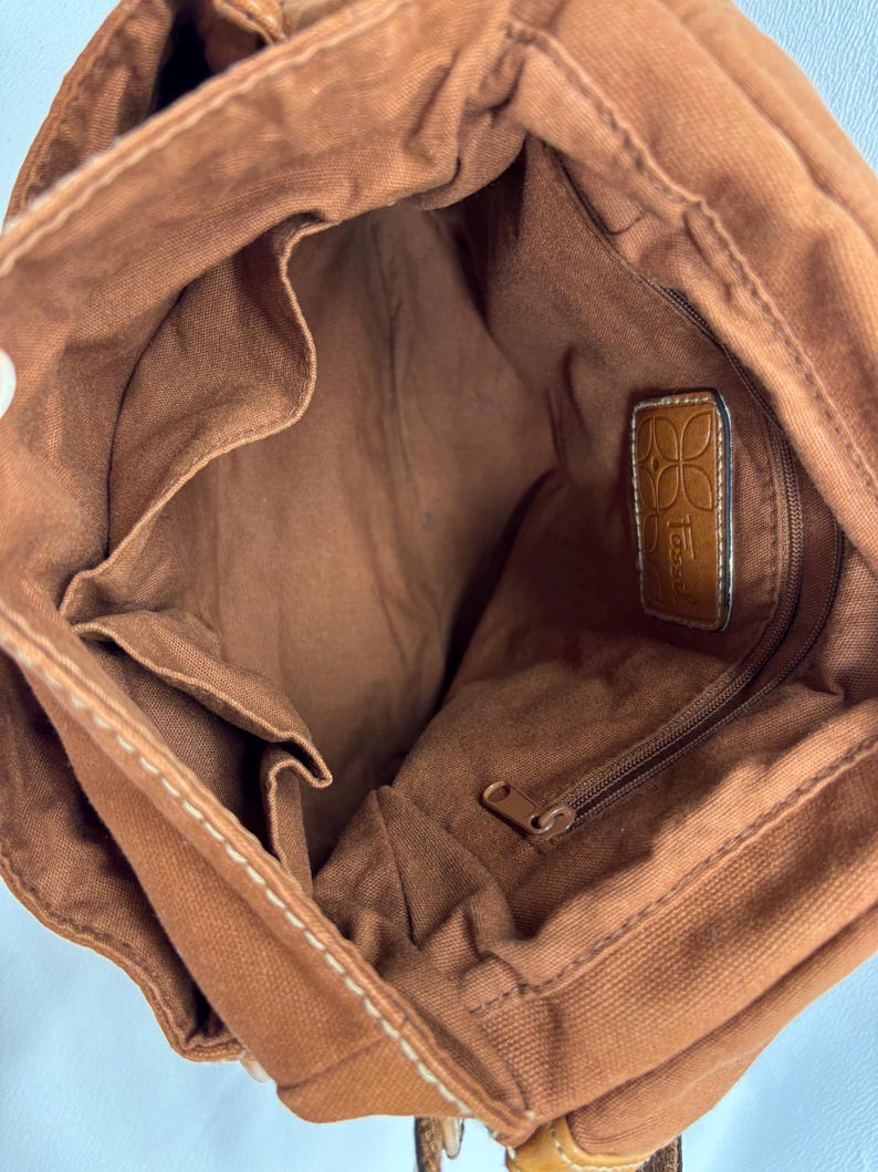 Pu&ograve; includere: Vista interna di una borsa tote in tela marrone con pi&ugrave; tasche interne e uno scomparto con cerniera. La borsa presenta cuciture bianche e un'etichetta in pelle con il nome del marchio. La fodera interna &egrave; di colore marrone abbinato.