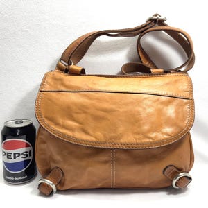 Pu&ograve; includere: Borsa a tracolla in pelle marrone chiaro con chiusura a patta e tracolla regolabile. La borsa ha una forma arrotondata e dettagli in metallo. Viene mostrata accanto a una lattina di Pepsi.