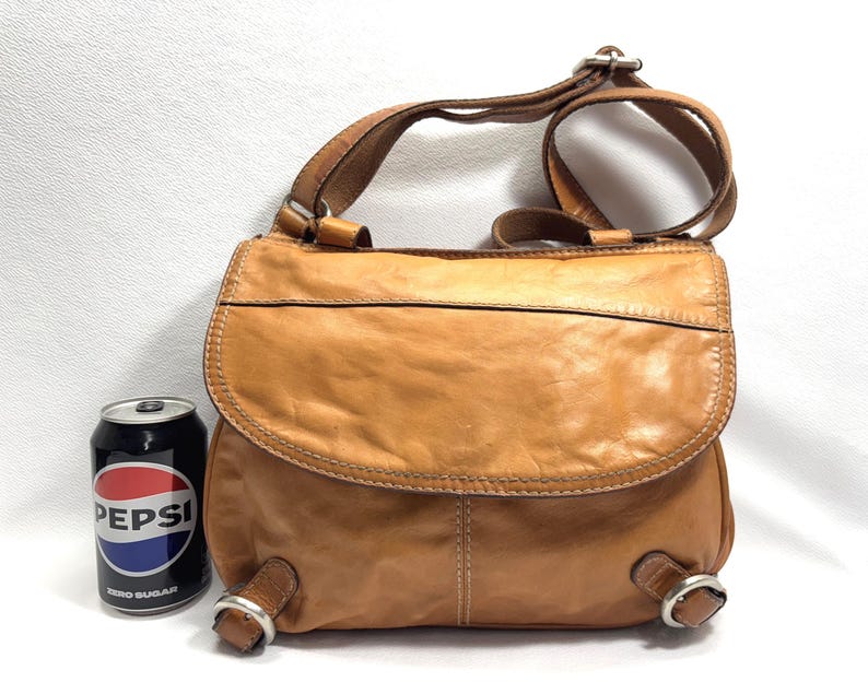 Pu&ograve; includere: Borsa a tracolla in pelle marrone chiaro con chiusura a patta e tracolla regolabile. La borsa ha una forma arrotondata e dettagli in metallo. Viene mostrata accanto a una lattina di Pepsi.