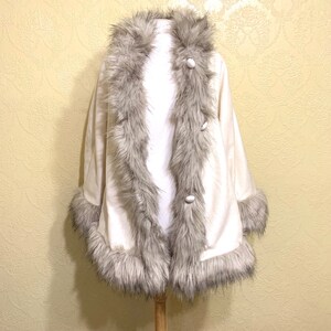 May include: A white faux fur cape with a gray faux fur trim around the collar, cuffs, and hem. The cape has a button closure.