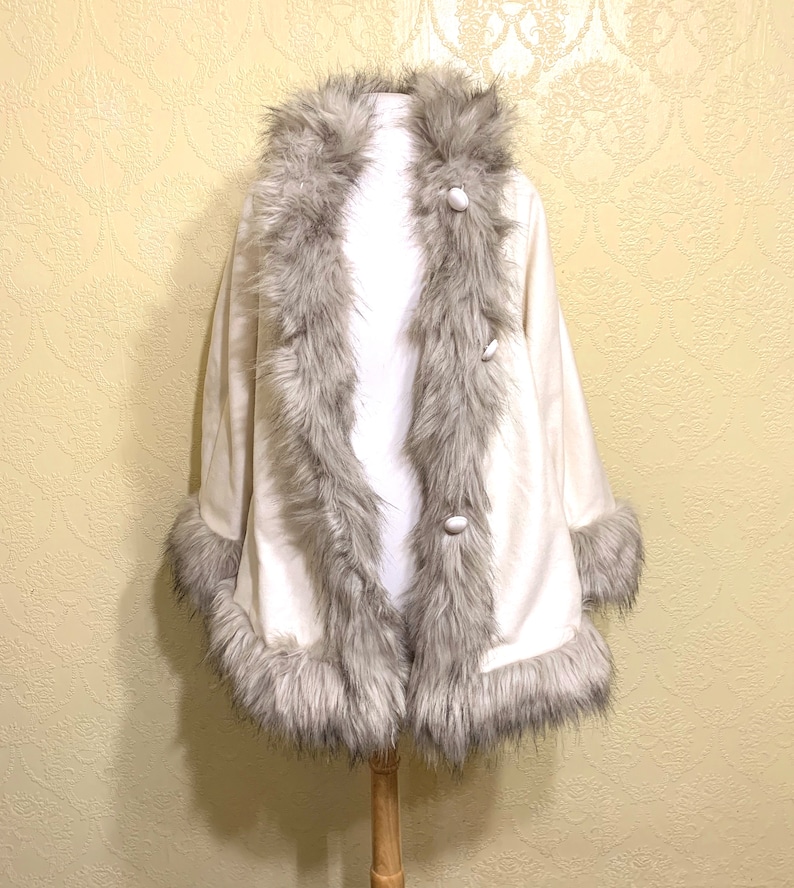 May include: A white faux fur cape with a gray faux fur trim around the collar, cuffs, and hem. The cape has a button closure.