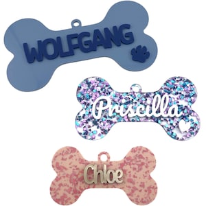 May include: Three dog bone-shaped pet tags. One is blue with the name "WOLFGANG" and a paw print. Another is glittery with the name "Priscilla". The third is pink with the name "Chloe". All have a small hole for attachment.